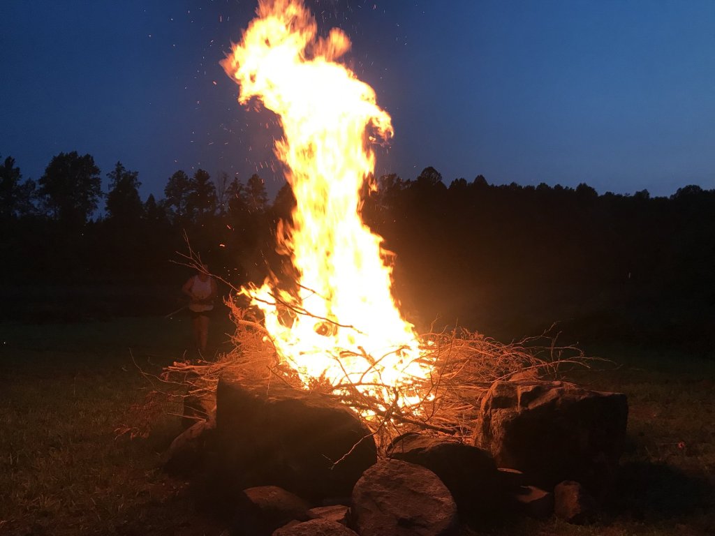 Tall bonfire burning against a dark treeline at night