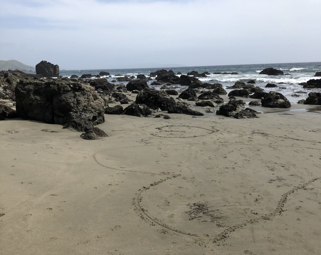 A heart drawn in beach sand among scattered rocks with the ocean behind